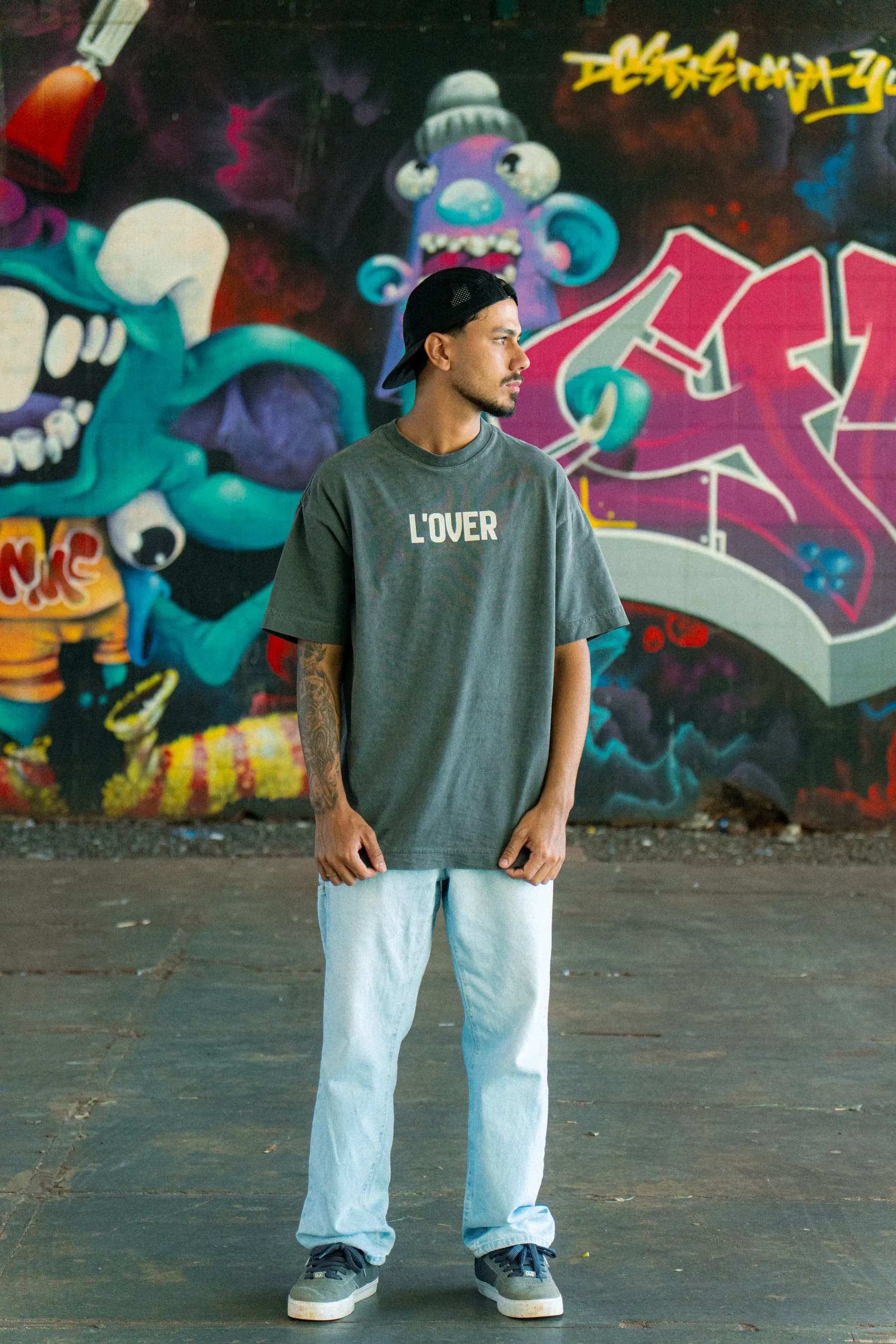 Modelo de pé com a camiseta concrete L'OVER diante de um muro grafitado.