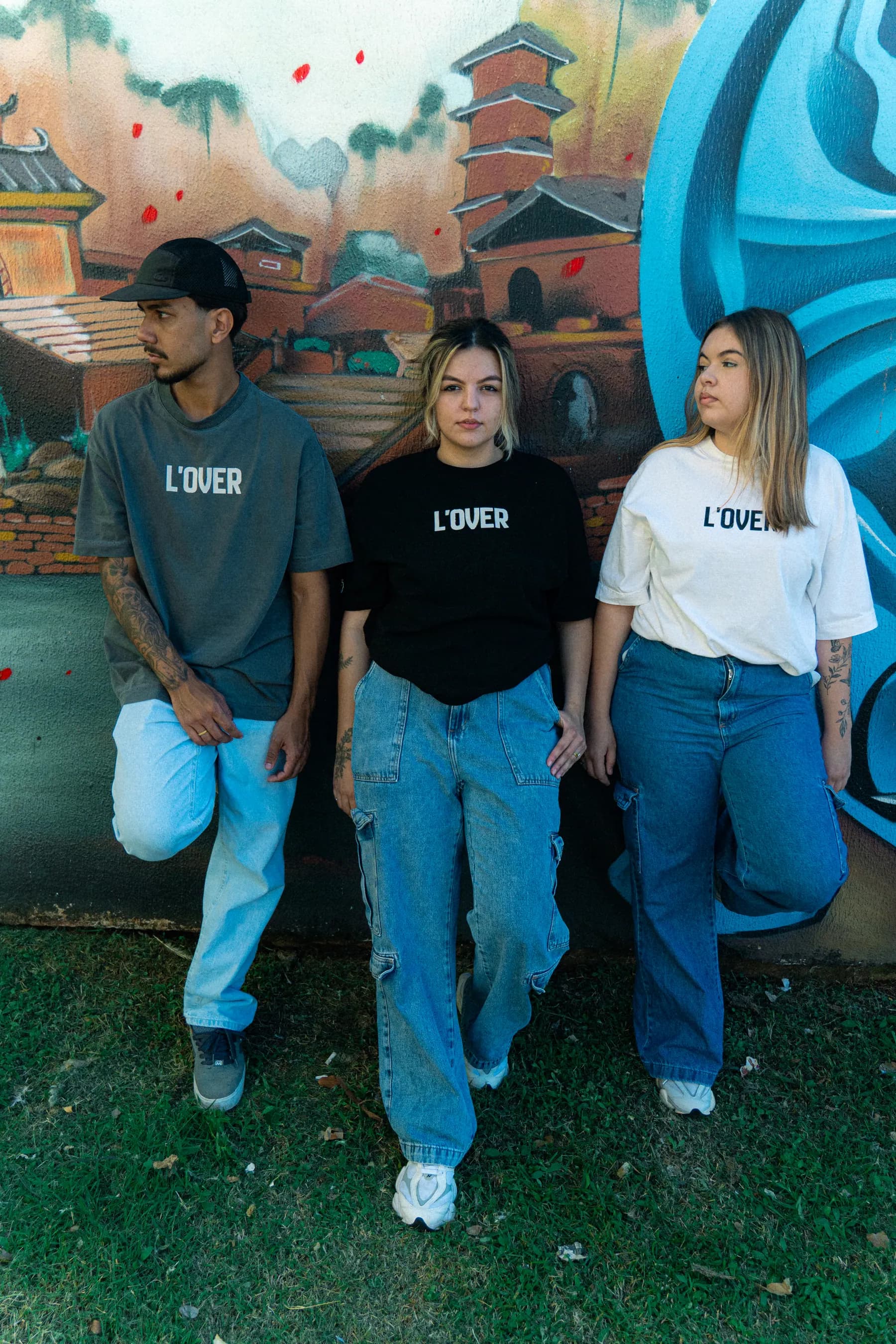 Três pessoas com camisetas L'OVER preta, off white e concrete diante de um mural grafitado.