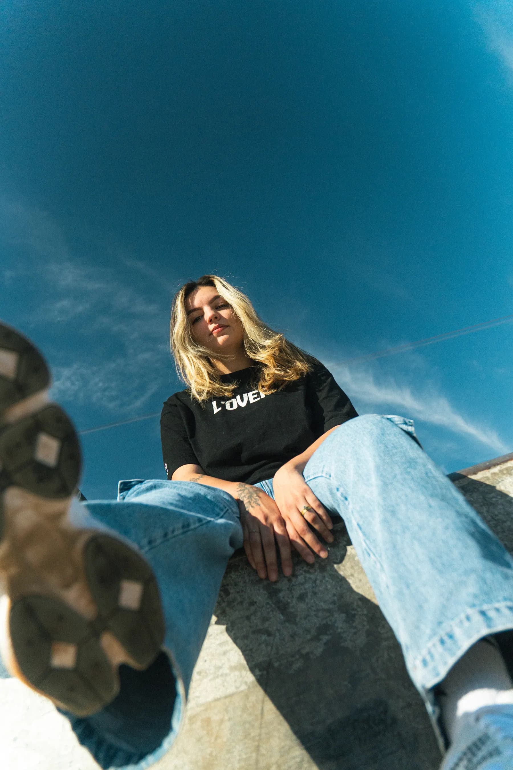 Modelo sentada em mureta de concreto, camiseta preta L'OVER, fotografada de baixo para cima contra o céu azul.