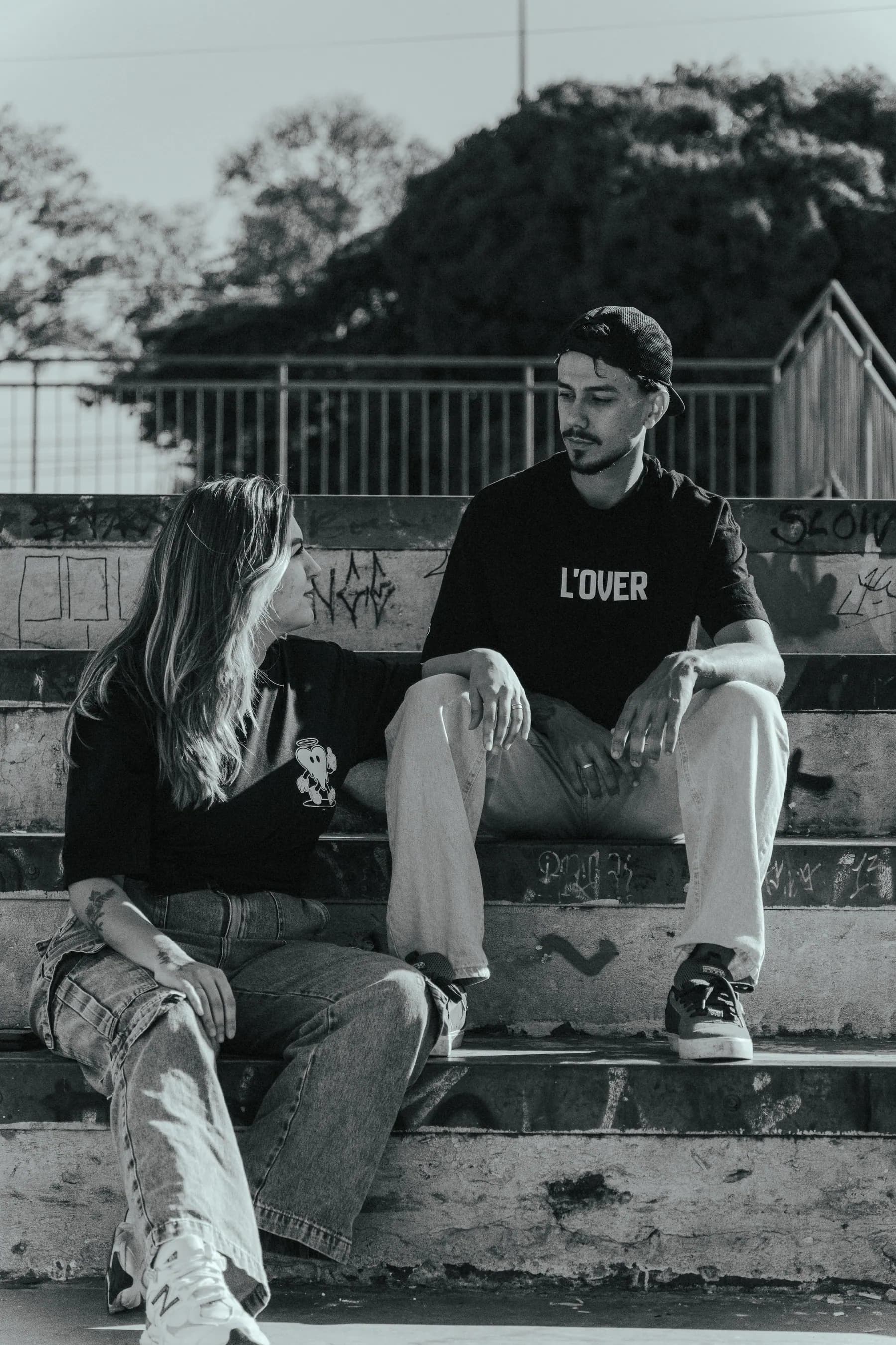 Dupla conversando na arquibancada do skatepark, camisetas L'OVER, fotografia em preto e branco.