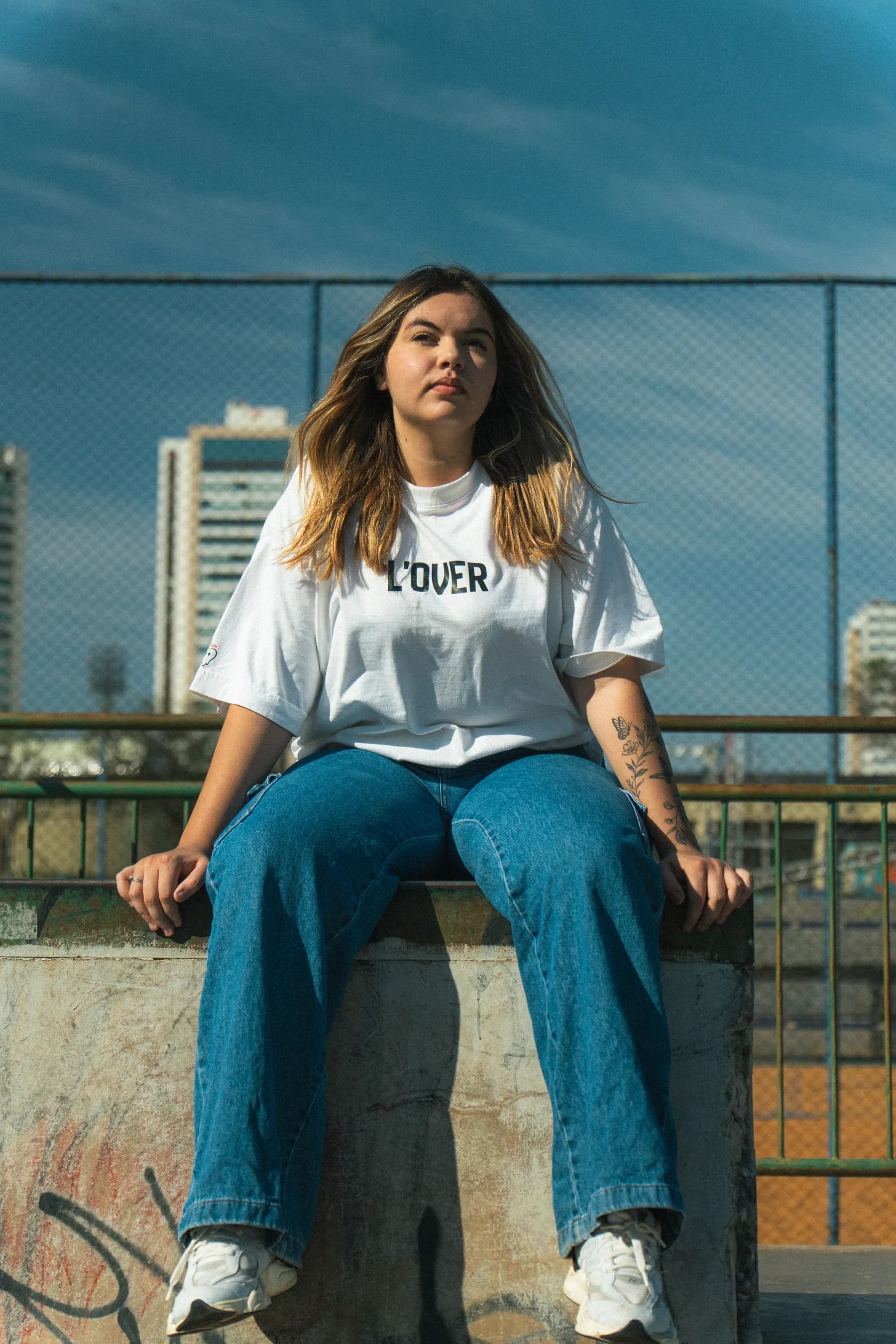 Modelo sentada com a camiseta off white L'OVER, arquibancada e céu ao fundo.