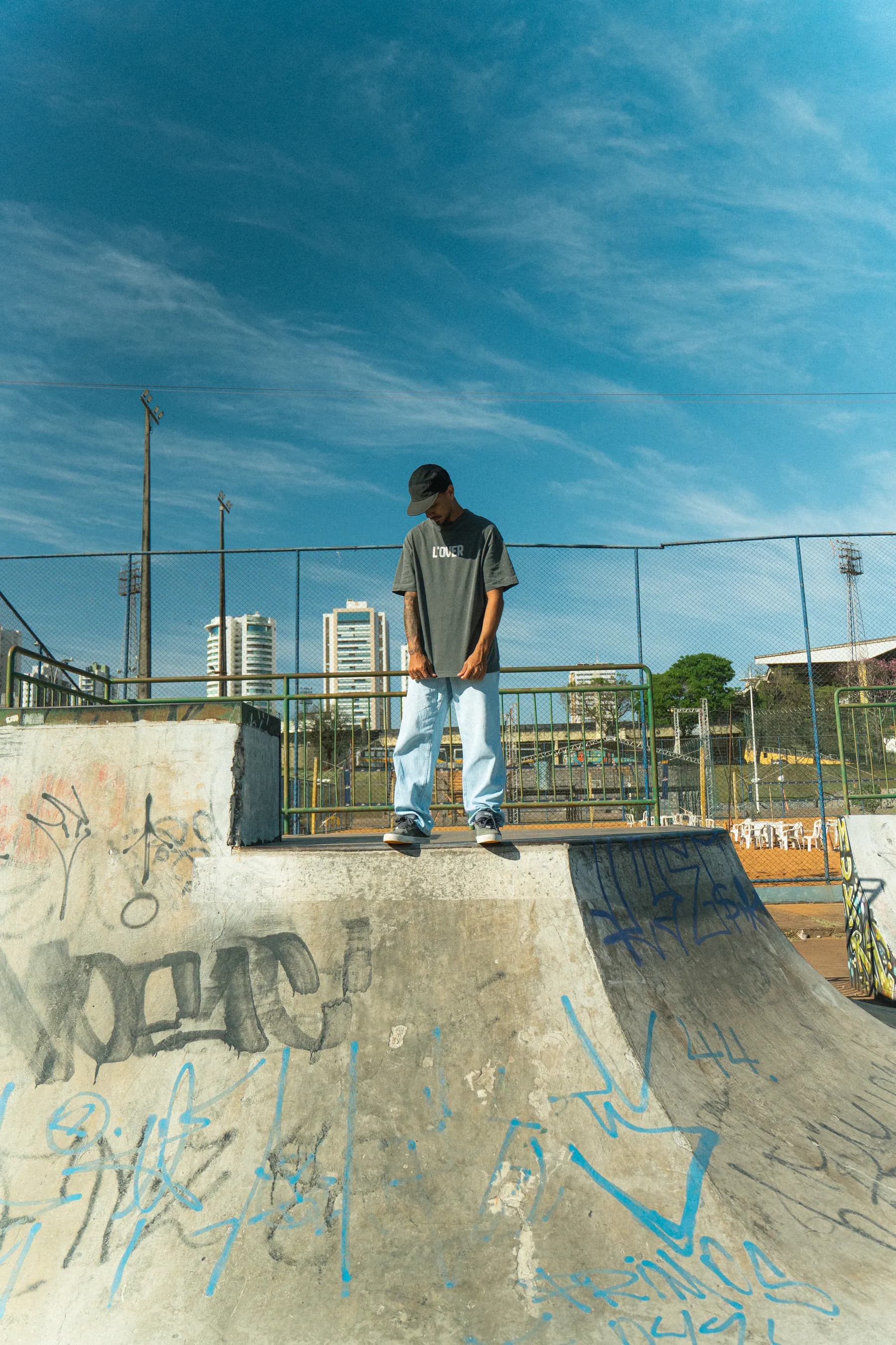 Modelo parado no topo da rampa do skatepark, céu aberto ao fundo.